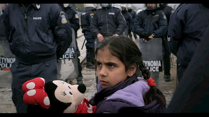 A young girl holds her stuffed Minnie Mouse as Greek police stand blocking the crossing into Macedonia. Thousands of refugees were stranded for months along this border in early 2016.